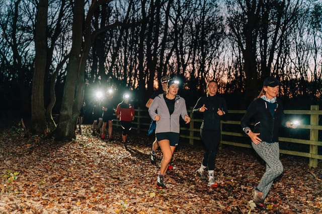 A group of people wearing headlamps running on a leaf-strewn path through a forest at dusk or dawn.